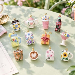 Könnte beinhalten: Eine Sammlung von elf Miniatur-Anhängern aus Keramik mit verschiedenen Motiven, darunter eine Teekanne, eine Sodaflasche, eine Kaffeetasse mit Latte Art, ein Blumenbecher, ein Geschenk, ein Einmachglas, ein Milchkarton und ein Teebeutel, mit Goldring.