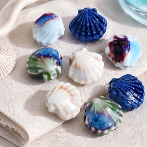 Könnte beinhalten: Eine Sammlung von Keramikperlen in Muschelform mit verschiedenen Glasuroberflächen, darunter einfarbiges Tiefblau, Weiß mit braunen Akzenten sowie mehrfarbige Mischungen aus Grün, Blau und Violett. Jede Perle hat ein sichtbares Loch zum Auffädeln.
