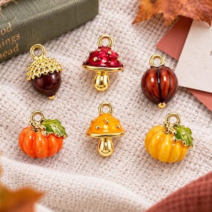 Può includere: Un set di sei ciondoli in metallo a tema autunnale, tra cui una ghianda marrone, un fungo rosso, una noce marrone, una zucca arancione con una foglia verde, un fungo giallo e una zucca gialla con una foglia verde, ciascuno con un anellino dorato.