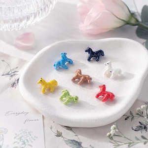 May include: A collection of seven small, glossy ceramic horse figurines in various colors including blue, dark blue, white, brown, yellow, green, and red, arranged on a white, irregular, textured ceramic dish with a matte finish.