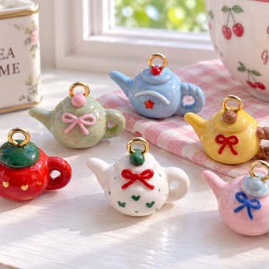 May include: A collection of six miniature ceramic teapot charms, each featuring a gold-tone metal loop at the top. The teapots are painted in various colors including red, green, blue, yellow, white, and pink, decorated with small bows and patterns.