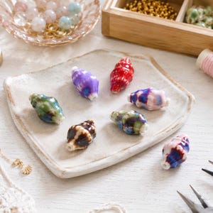May include: A collection of seven ceramic beads shaped like small seashells, each featuring a unique glossy glaze in colors including purple, red, green, brown, and blue, arranged on a square, off-white, handmade ceramic tray.