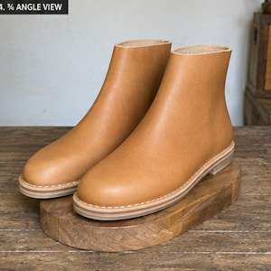 May include: A pair of tan leather ankle boots with a smooth finish, featuring a rounded toe, a simple slip-on design, and a layered light-coloured sole with visible white stitching around the base. The boots have a clean, unadorned exterior.