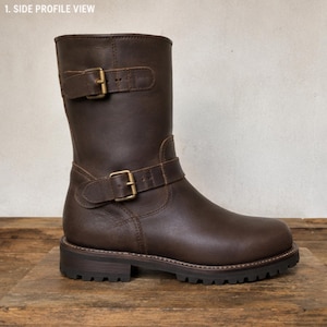 May include: A dark brown leather mid-calf boot featuring two adjustable straps with antique brass-toned metal buckles, one positioned at the top of the shaft and one at the ankle, set on a thick, dark brown lugged rubber sole with visible stitching.