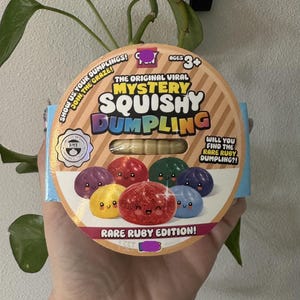 May include: A round cardboard packaging for a Mystery Squishy Dumpling toy. The front features text including The Original Viral Mystery Squishy Dumpling, Rare Ruby Edition, and illustrations of colorful, smiling dumpling characters in various shades.