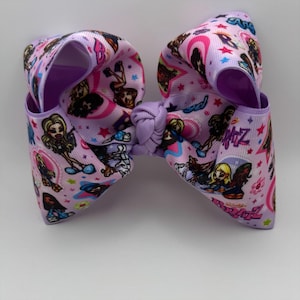 May include: A large hair bow made of light purple grosgrain ribbon featuring a repeating pattern of Bratz doll characters, pink and blue stars, and the Bratz logo in purple and black. The center is gathered with a matching purple ribbon knot.