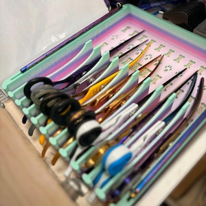 Op de afbeelding: Een licht mintgroen organisator-dienblad met een pastelkleurige regenboogrand en een pootafdrukpatroon op de bodem. Het dienblad bevat meerdere paren professionele trimschaar in diverse kleuren, waaronder goud, paars en blauw, met zwarte vingerringen.