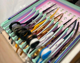 Puede incluir: Un estuche de almacenamiento multicolor con un marco verde claro y una base interior de rayas pastel con un patrón repetitivo de pequeños huesos y huellas de patas. El estuche contiene varios pares de tijeras de aseo con mangos variados.