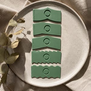 May include: Five sage green clay scrapers arranged vertically, each featuring a circular indentation and unique edge designs including straight, angled, chevron, serrated, and wavy patterns, with the text Pottery Supply Co. embossed on each.