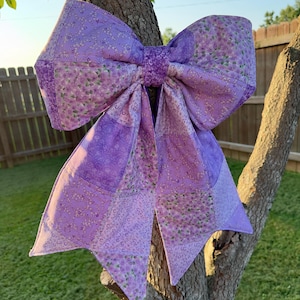 May include: A large decorative bow constructed from quilted fabric featuring a patchwork design in various shades of purple. The fabric displays multiple floral and geometric patterns with subtle glitter accents throughout the surface.
