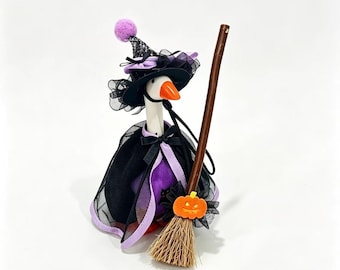 May include: A white goose figurine dressed as a witch, wearing a black and purple hat with a lavender pom-pom and a black cape with purple trim. The goose holds a brown wooden broomstick featuring a small orange jack-o'-lantern decoration.