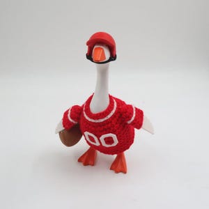 May include: A white goose figurine wearing a red crochet jersey with white trim and the number 00 on the front. The goose also wears a red plastic helmet with a black chin strap and holds a small brown football under its right wing.