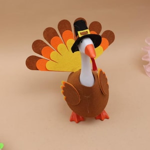 May include: A decorative goose figurine dressed as a turkey, featuring a white neck, orange beak, and brown felt body. It wears a black pilgrim hat with a yellow buckle and has a large, fanned tail made of layered brown, orange, and yellow felt feathers.