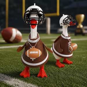 May include: Two white duck figurines wearing miniature white football helmets with black face masks and brown football jerseys with white stripes. Each duck wears a silver ball chain necklace featuring a small brown football pendant.