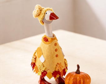 May include: A white goose figurine wearing a yellow knit hat and a yellow felt coat decorated with orange and red fabric autumn leaves. The goose has a dark orange beak and feet. Beside the goose sits a small, round, orange pumpkin figurine.