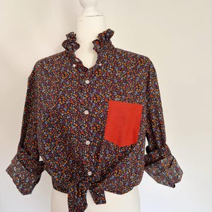 May include: A long-sleeved button-down shirt featuring a dark navy blue base with a dense multicolored floral pattern. The shirt includes a ruffled collar, a solid orange chest pocket, and a tie-front hem. The sleeves are rolled up at the cuffs.