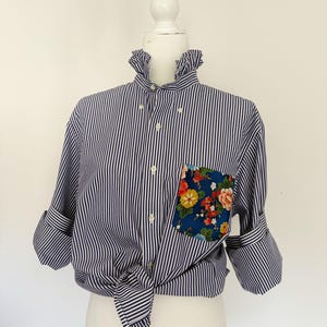 May include: A navy blue and white vertical striped button-down shirt featuring a ruffled collar and a single chest pocket with a blue floral pattern. The shirt has long sleeves with rolled cuffs and is tied at the waist, displayed on a mannequin.