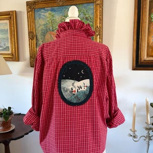 May include: A red and white checkered button-down shirt with a ruffled collar and long sleeves. The back features a large, oval-shaped embroidered patch depicting a nighttime winter landscape with a small house, trees, and a crescent moon.