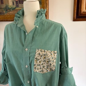 May include: A green and white gingham button-down shirt featuring a ruffled collar and a single chest pocket with a white floral print. The shirt has long sleeves with rolled cuffs and a front button closure, displayed on a white mannequin torso.
