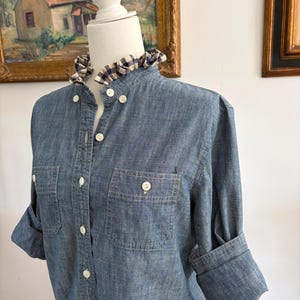 May include: A blue chambray button-down shirt featuring a ruffled collar with a navy and white gingham check pattern. The shirt includes long sleeves with rolled cuffs, two chest pockets with button closures, and white buttons down the front.