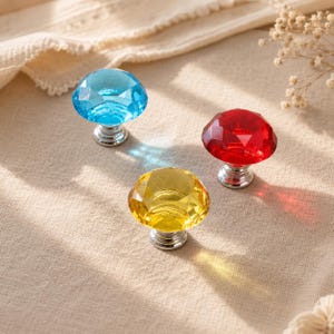 May include: Three faceted cabinet knobs with a diamond-like shape, featuring one blue, one red, and one yellow translucent glass top. Each knob is attached to a circular silver-toned metal base, creating a reflective and colorful hardware set.
