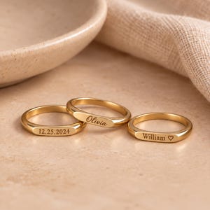 Könnte beinhalten: Drei goldfarbene Metallringe mit flachen, polierten Oberflächen. Ein Ring ist mit dem Datum 12.25.2024 graviert, ein weiterer zeigt den Namen Olivia in Schreibschrift und der dritte den Namen William, gefolgt von einem kleinen Herzsymbol.