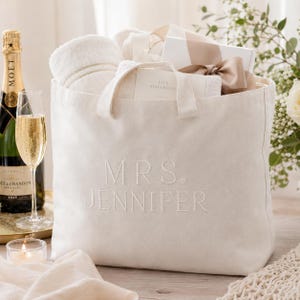Pode incluir: Uma bolsa tote de lona na cor creme com o texto bordado MRS. JENNIFER na frente. A bolsa está preenchida com uma toalha branca, uma caixa de presente branca com uma fita bege e um pequeno cartão com o texto LIVE BEAUTIFULLY.