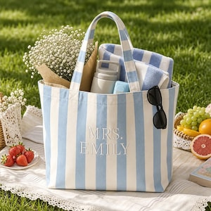 May include: A light blue and white vertical striped canvas tote bag featuring the embroidered text MRS. EMILY on the front. The bag contains a white towel, a light gray insulated bottle, and a bouquet of baby's breath flowers.