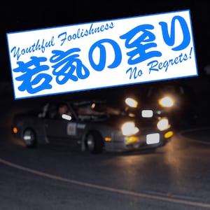 Könnte beinhalten: Ein rechteckiger Aufkleber mit leuchtend blauem Text auf weißem Hintergrund. Der Text lautet Youthful Foolishness, No Regrets und japanische Schriftzeichen. Unter dem Aufkleber ist ein unscharfes, dunkles Bild eines Sportwagens bei Nacht.