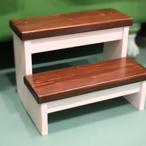 May include: A two-step stool featuring a white painted wooden frame and two dark brown stained wooden steps. The stool has a rectangular design with a lower step and a higher step, showcasing a clear wood grain texture on the surfaces.