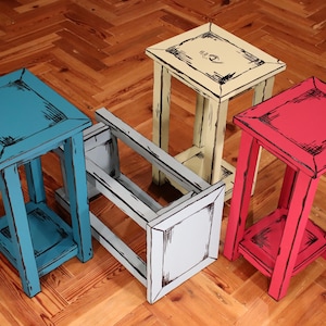 Puede incluir: Cuatro mesas auxiliares de madera en colores turquesa, crema, gris claro y rosa brillante. Cada mesa tiene una superficie cuadrada, cuatro patas y un estante inferior, con contornos pintados en negro que crean un efecto visual en 2D.