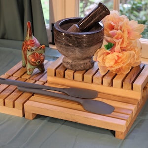 May include: A two-tiered wooden slat display stand holding a dark grey stone mortar and pestle, a small painted ceramic cat figurine, two grey silicone spatulas, and a cluster of light peach-colored artificial flowers arranged on the top tier.