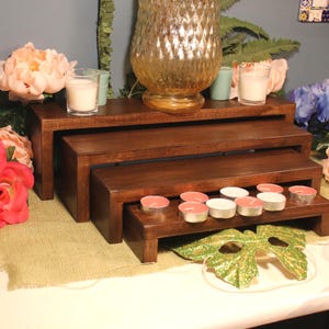 May include: A set of three tiered rectangular wooden display risers with a dark brown finish. The risers are arranged in a cascading formation, holding small white candles in glass holders and several pink and blue artificial flowers.