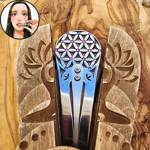 May include: A silver-toned metal jaw harp featuring a circular head with a geometric flower of life pattern inlaid with small purple and blue crystals. The instrument has two visible screws and a central metal tongue, presented within a wooden holder.
