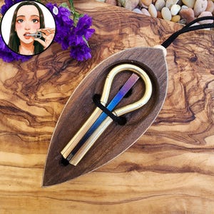 Könnte beinhalten: Eine Maultrommel aus Messing mit einer zentralen, irisierend blau und violett schimmernden Metallzunge, befestigt mit einer schwarzen Kordel in einem tropfenförmigen Rahmen aus dunklem Holz. Eine Illustration zeigt die Spielweise.