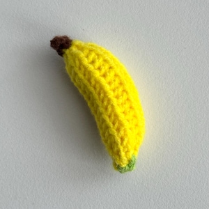 Könnte beinhalten: Eine kleine gehäkelte Banane mit einem leuchtend gelben Körper, einem dunkelbraunen Stiel an der Oberseite und einer hellgrünen Spitze an der Unterseite. Die Garnstruktur ist auf der gesamten geschwungenen Form dieses handgefertigten Objekts sichtbar.