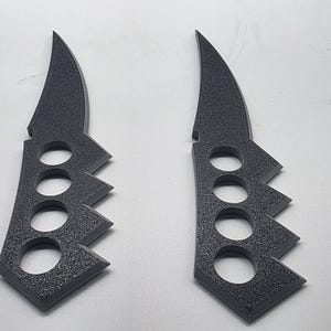 Puede incluir: Dos objetos de color gris oscuro, texturizados y de perfil plano con forma de cuchillo y mangos integrados tipo pu&ntilde;o americano. Cada pieza presenta una secci&oacute;n de hoja curva y un mango con cuatro orificios circulares para los dedos.