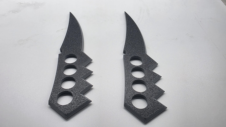 Puede incluir: Dos objetos de color gris oscuro, texturizados y de perfil plano con forma de cuchillo y mangos integrados tipo pu&ntilde;o americano. Cada pieza presenta una secci&oacute;n de hoja curva y un mango con cuatro orificios circulares para los dedos.