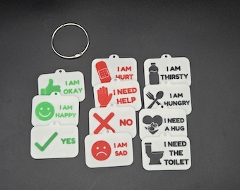Puede incluir: Un conjunto de tarjetas de comunicación rectangulares blancas con iconos y texto en negro, rojo y verde, incluyendo I AM OKAY, I AM HAPPY, YES, I AM HURT, I NEED HELP, NO, I AM SAD, I AM THIRSTY, I AM HUNGRY, I NEED A HUG y I NEED THE TOILET, con una anilla metálica.