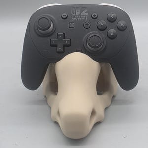 May include: A black Nintendo Switch Pro controller resting on a beige, skull-shaped display stand. The controller features a central Nintendo Switch logo, two analog sticks, a directional pad, and standard action buttons labeled A, B, X, and Y.