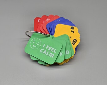Puede incluir: Un conjunto de tarjetas rectangulares de plástico en rojo, azul, amarillo y verde, unidas por una anilla metálica. Cada tarjeta presenta un icono y texto en relieve, como la tarjeta verde con una cara sonriente y el texto I FEEL CALM.