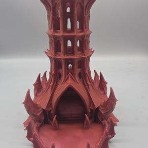 May include: A matte reddish-brown 3D printed dice tower featuring a tall, multi-tiered structure with pointed, arched windows and a wide, open base. The exterior surface displays intricate, sharp, wing-like protrusions and vertical columns.