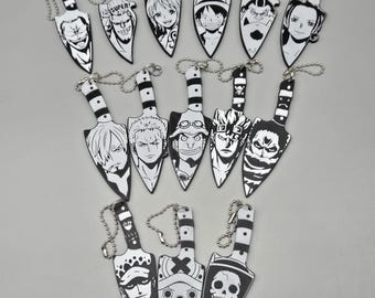 Puede incluir: Una colección de catorce llaveros en forma de daga en blanco y negro, cada uno con una ilustración de personaje única de la serie One Piece. Cada llavero incluye una cadena de bolas de metal unida al mango, mostrando un detallado arte lineal.