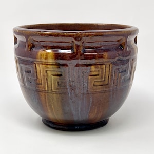 May include: A round ceramic bowl featuring a glossy brown glaze with vertical streaks of lighter tan and blue. The exterior surface is decorated with a raised, repeating Greek key pattern that encircles the upper portion of the vessel.