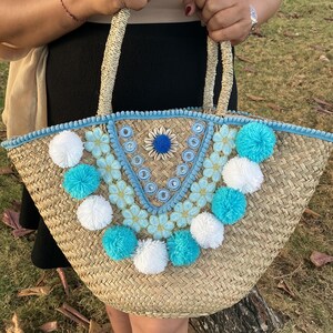 May include: A woven straw tote bag featuring a V-shaped decorative panel adorned with light blue floral lace trim, small circular mirrors, and a central shell flower. The bag is embellished with alternating white and turquoise pom-poms.