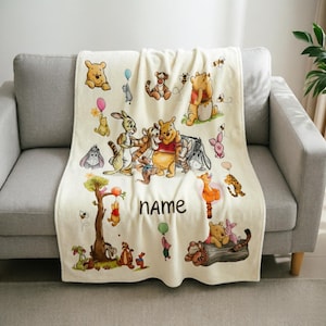 May include: A cream-coloured fleece blanket featuring a repeating pattern of Winnie the Pooh characters including Piglet, Tigger, and Eeyore. The design includes colourful balloons, bees, and trees, with the word NAME printed in the centre in black.