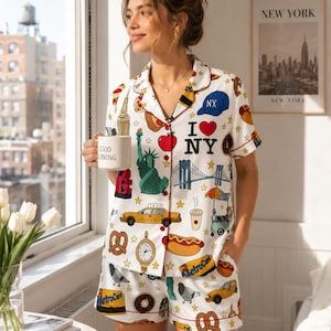 May include: A white short-sleeve button-down pajama shirt and matching shorts set featuring a colorful New York City print. The pattern includes the Statue of Liberty, yellow taxis, hot dogs, pretzels, and the text I LOVE NY with red piping.