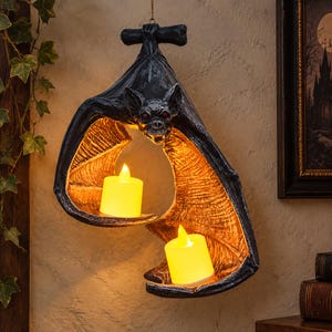 May include: A hanging bat-shaped candle holder in a dark charcoal finish with textured wings that cradle two glowing yellow LED pillar candles. The bat features detailed facial features and red eyes, suspended by a thin chain for display.