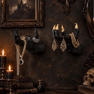 May include: Two wall-mounted black hand sculptures with gold-painted fingernails. The left hand holds a gold chain necklace with a black pendant and several rings. The right hand displays a pair of gold filigree earrings and a beaded gold bracelet.