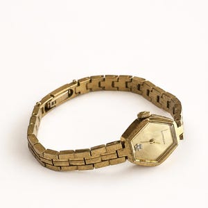 May include: A gold-toned wristwatch featuring a hexagonal case and a matching link bracelet. The watch face is gold with a single small clear stone accent at the bottom and the brand name Wittnauer printed in black on the right side.